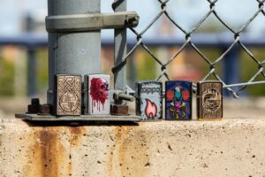 Lifestyle image of Eagle, Snake, Skull Design Iridescent Windproof Lighter standing with four other lighters in front of a chain link fence
